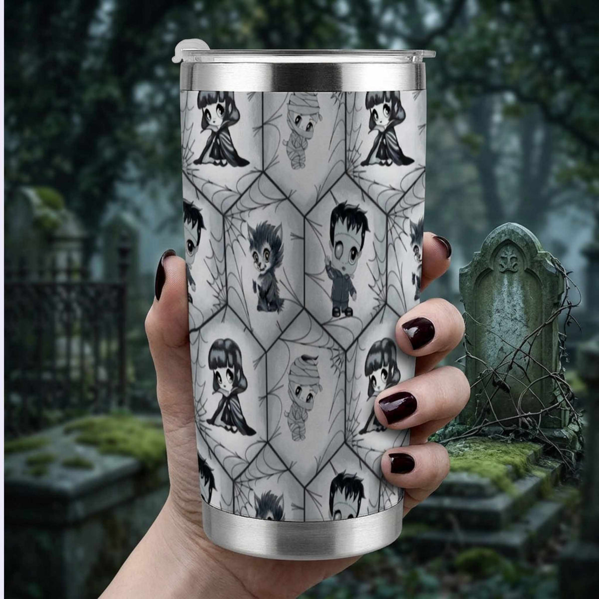 Hand with dark burgundy nails holding the chibi monster tumbler in front of a misty cemetery backdrop
