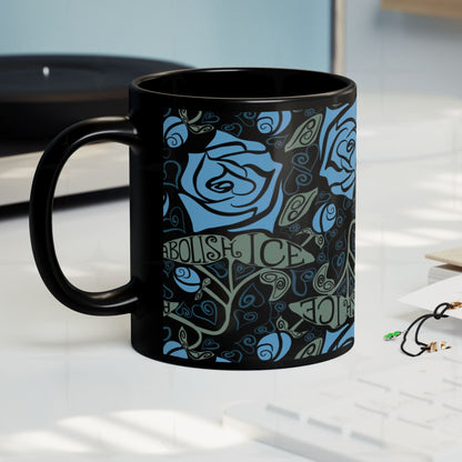 Abolish ICE black ceramic mug on a desk surface, gothic blue rose and hand-lettered activist wrap print, side view