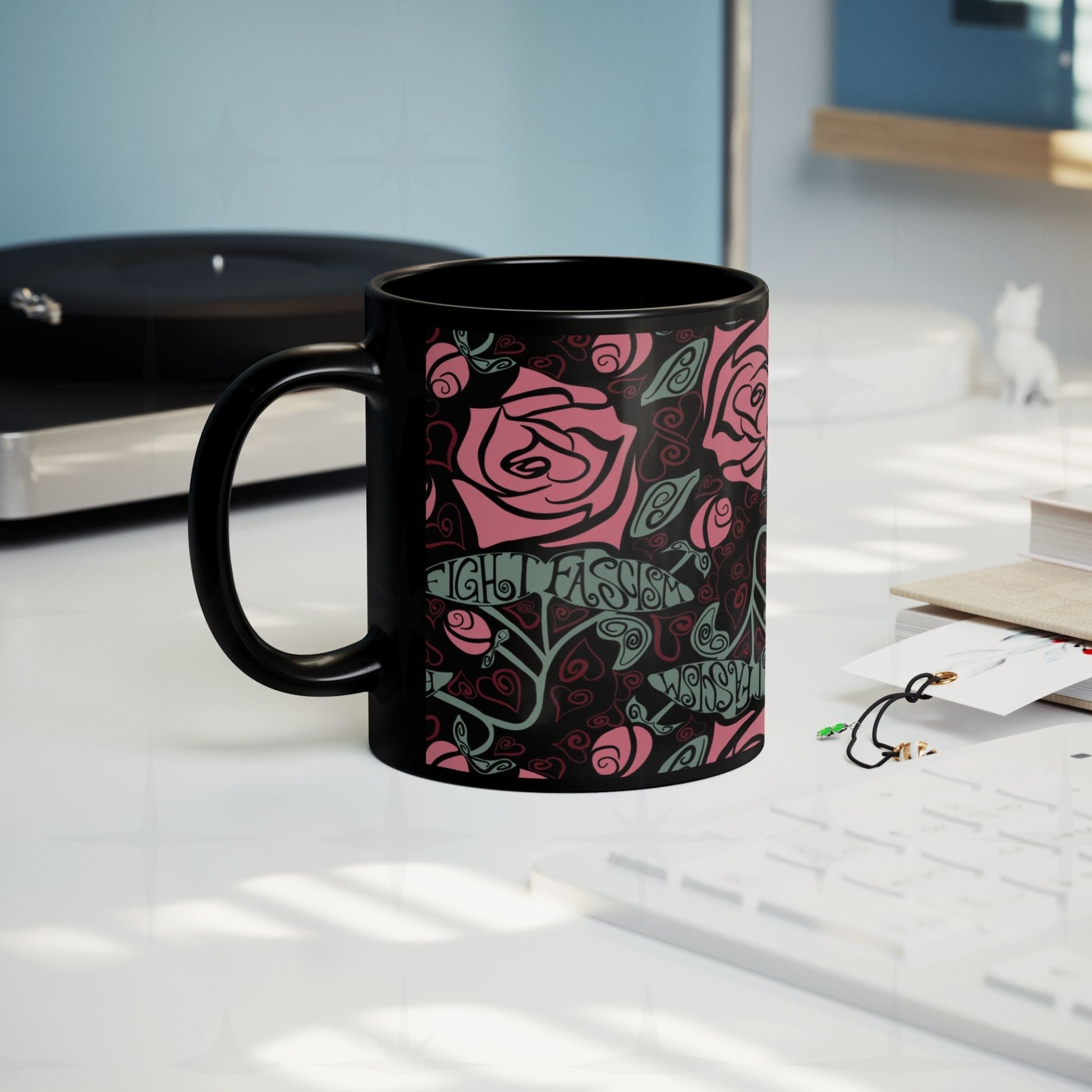 Fight Fascism anti-fascist coffee mug on a desk beside a record player, Art Nouveau rose pattern visible, cozy creative workspace lifestyle shot