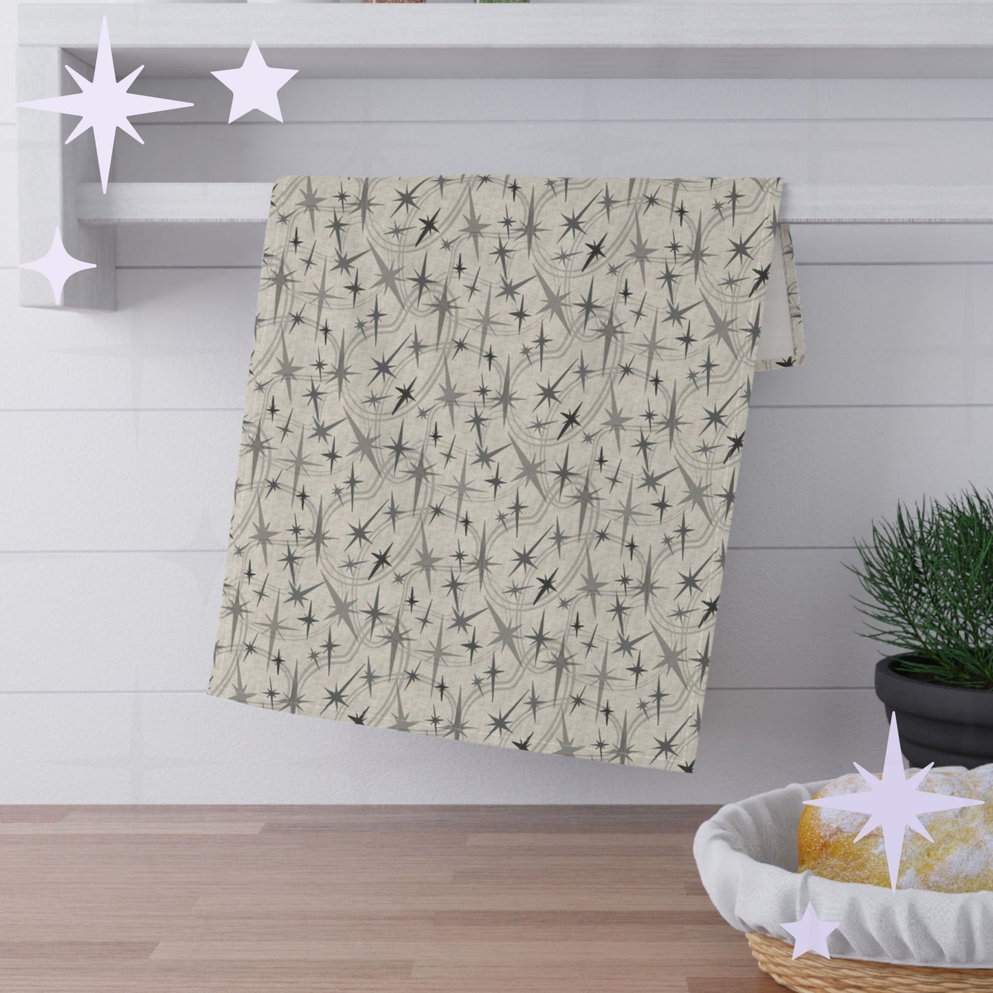 Warm gray atomic starburst cotton twill kitchen towel hanging from a round chrome hook against bright white subway tile with wooden shelves and stacked plates visible