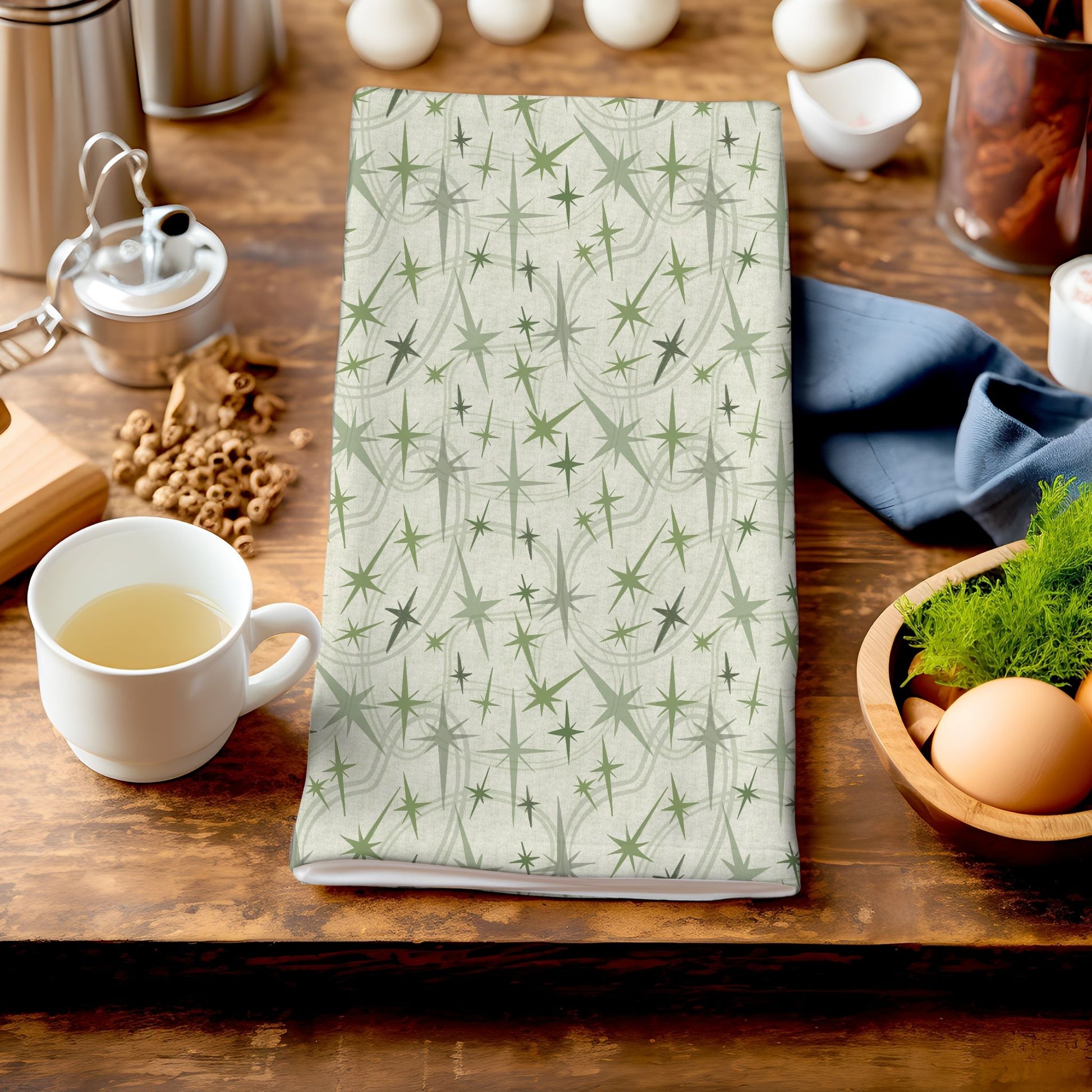 Sage green atomic starburst tea towel folded on a dark wood kitchen counter beside a white ceramic mug of tea, a bowl of eggs, and dried botanicals in the background