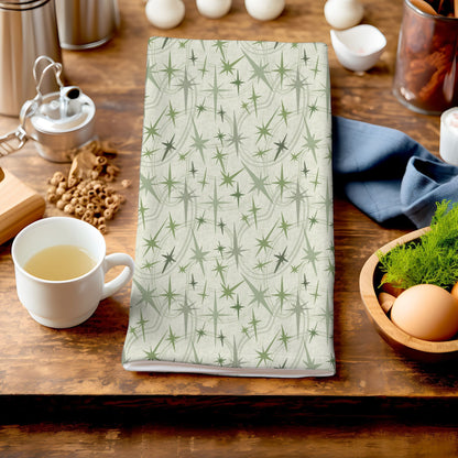 Sage green atomic starburst tea towel folded on a dark wood kitchen counter beside a white ceramic mug of tea, a bowl of eggs, and dried botanicals in the background