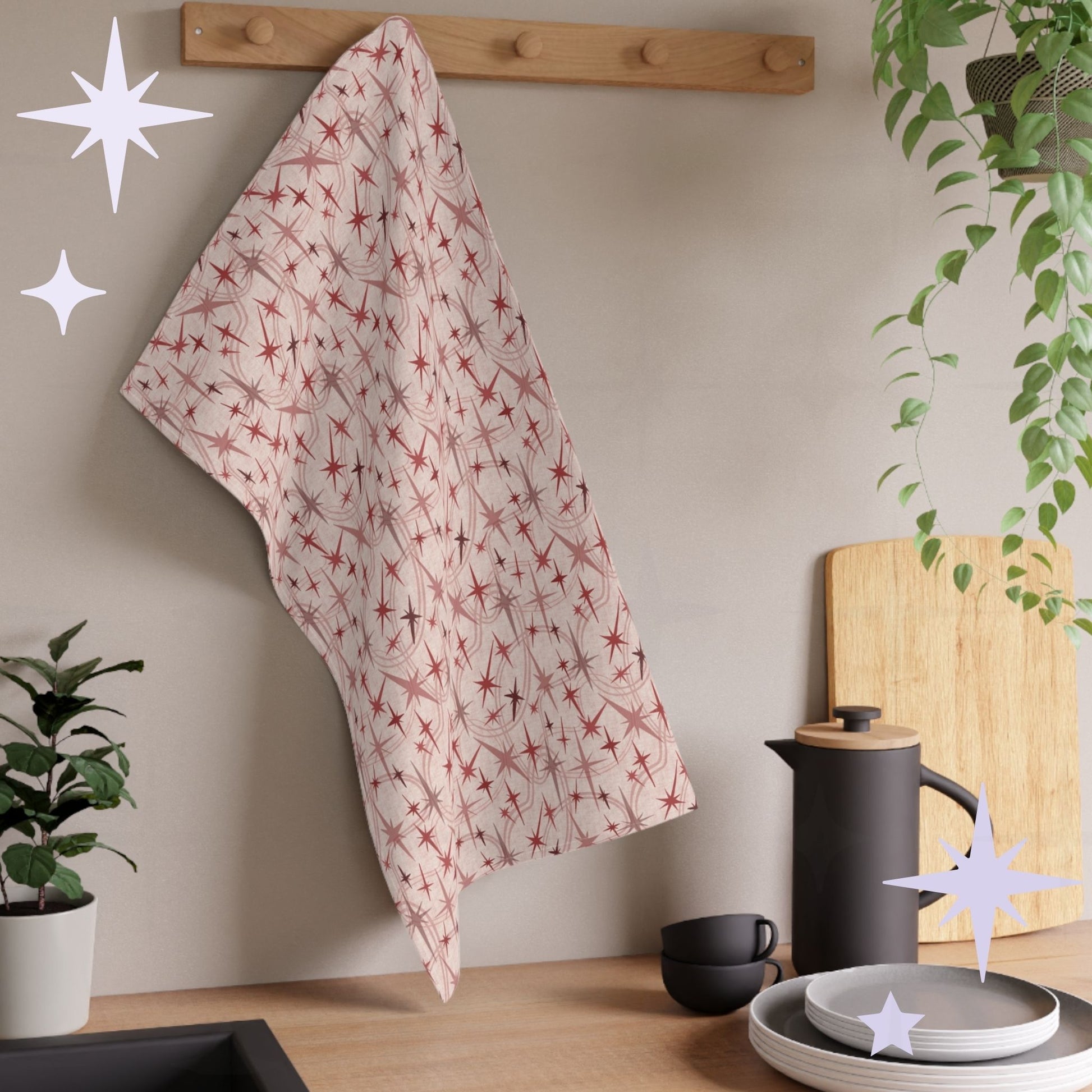 Dusty rose atomic starburst tea towel hanging from a wooden peg rail in a warm modern kitchen with houseplants, matte black coffee press, cutting board, and stacked gray plates