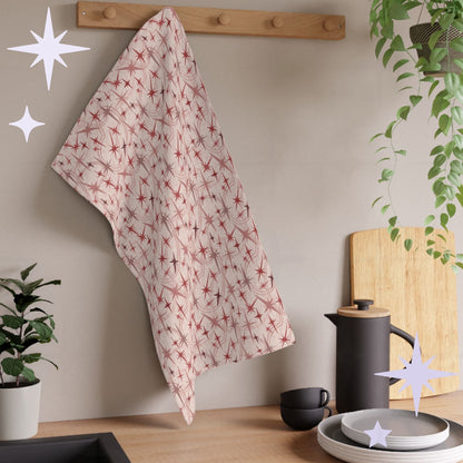 Dusty rose atomic starburst tea towel hanging from a wooden peg rail in a warm modern kitchen with houseplants, matte black coffee press, cutting board, and stacked gray plates