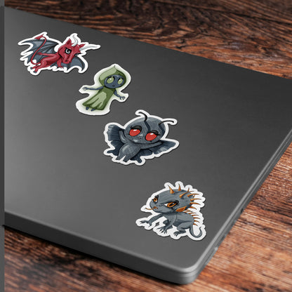 Four cryptid stickers applied to the corner of a gray laptop lid on a wood desk