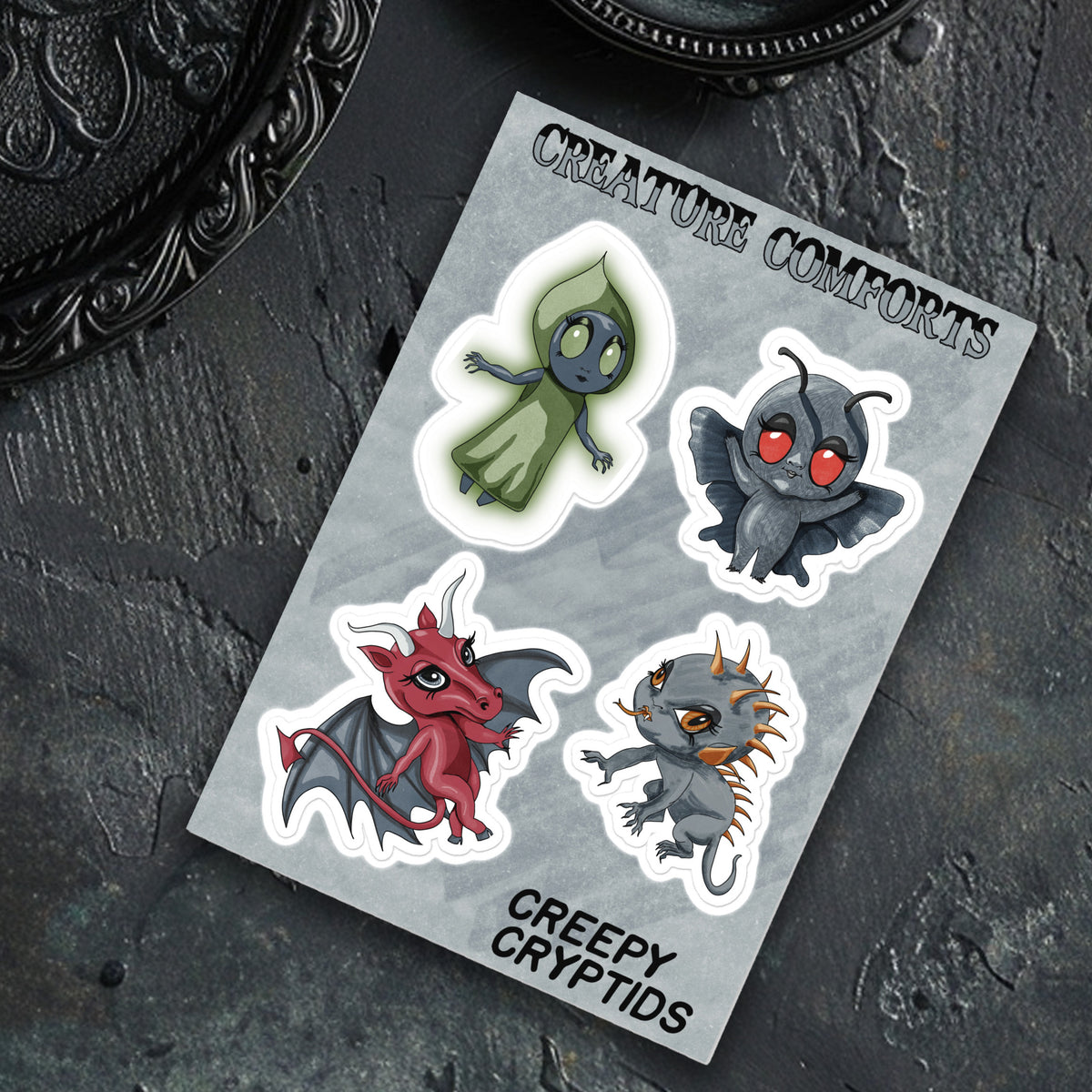 Cryptid sticker sheet on a slate surface next to a scrolled black metal frame