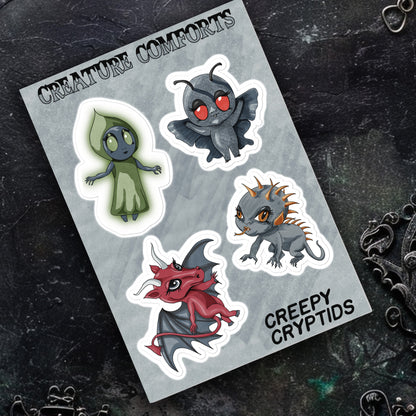 Creepy Cryptids sticker sheet resting on dark textured stone beside ornate silver filigree jewelry