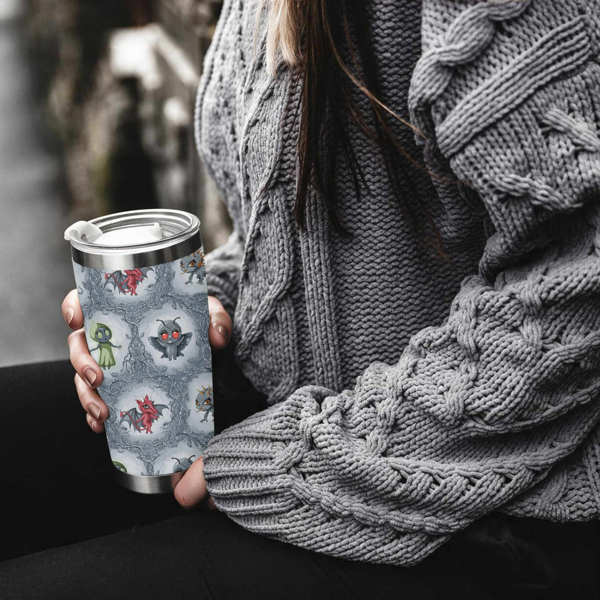 Woman in a chunky gray cable-knit sweater holding the colorful cryptid tumbler on a city street