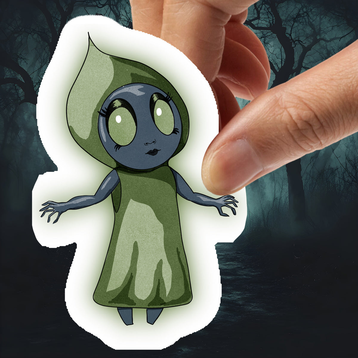 Hand holding the Flatwoods Monster sticker, a gray figure in a green hood with large pale eyes, against dark woods