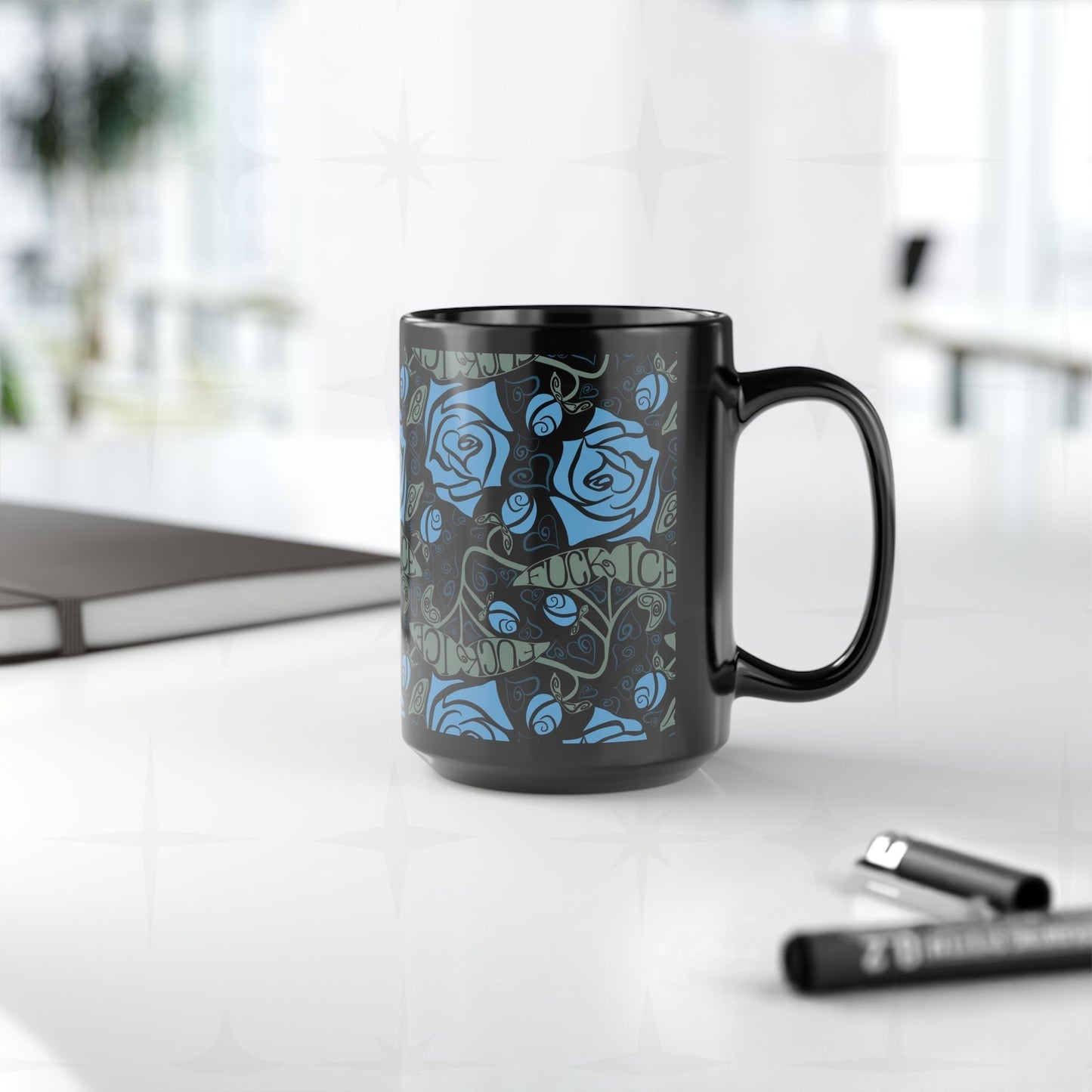 Dark floral hidden message activist mug sitting on a white desk beside a laptop and markers, showing the botanical rose pattern and concealed anti-ICE lettering in sage green on a 15oz glossy black ceramic mug