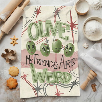 Funny cotton tea towel with olive characters and hand-lettered pun laid among baking ingredients and rolling pins on marble surface