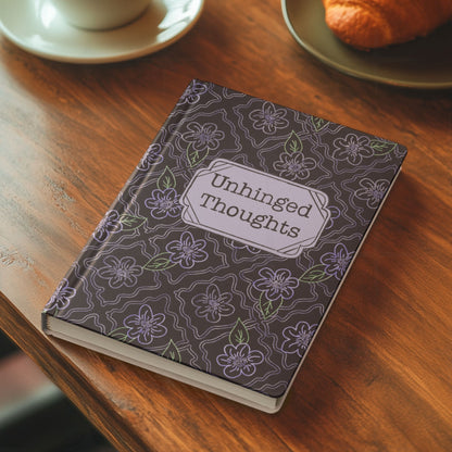 Funny floral hardcover journal on wooden café table beside a cup of coffee and croissant showing Unhinged Thoughts lavender cover design