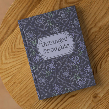 Funny floral hardcover journal with Unhinged Thoughts cover lying on round wooden surface, angled view showing charcoal and lavender ogee pattern