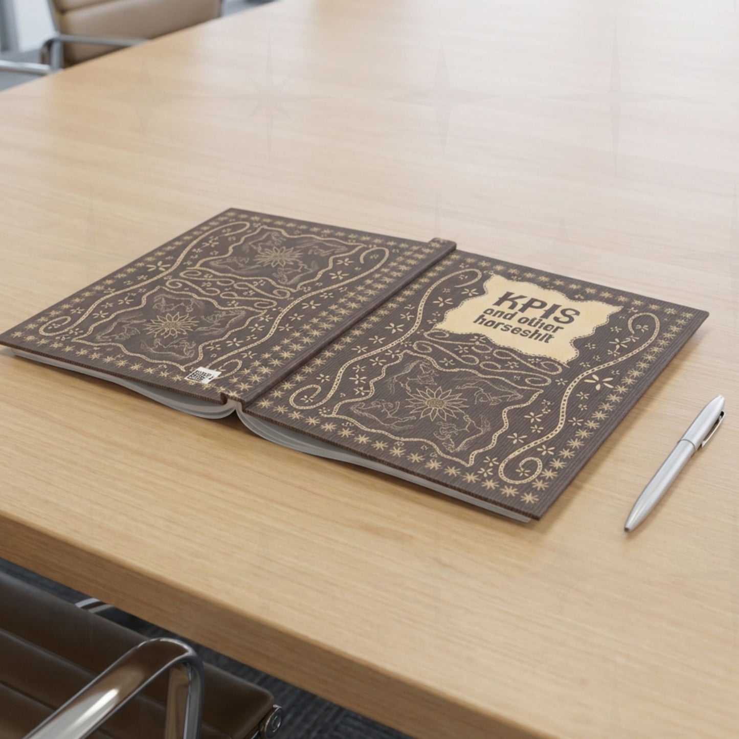 KPI profanity journal lying open flat on conference table beside silver pen showing both covers with vintage equestrian embroidered design