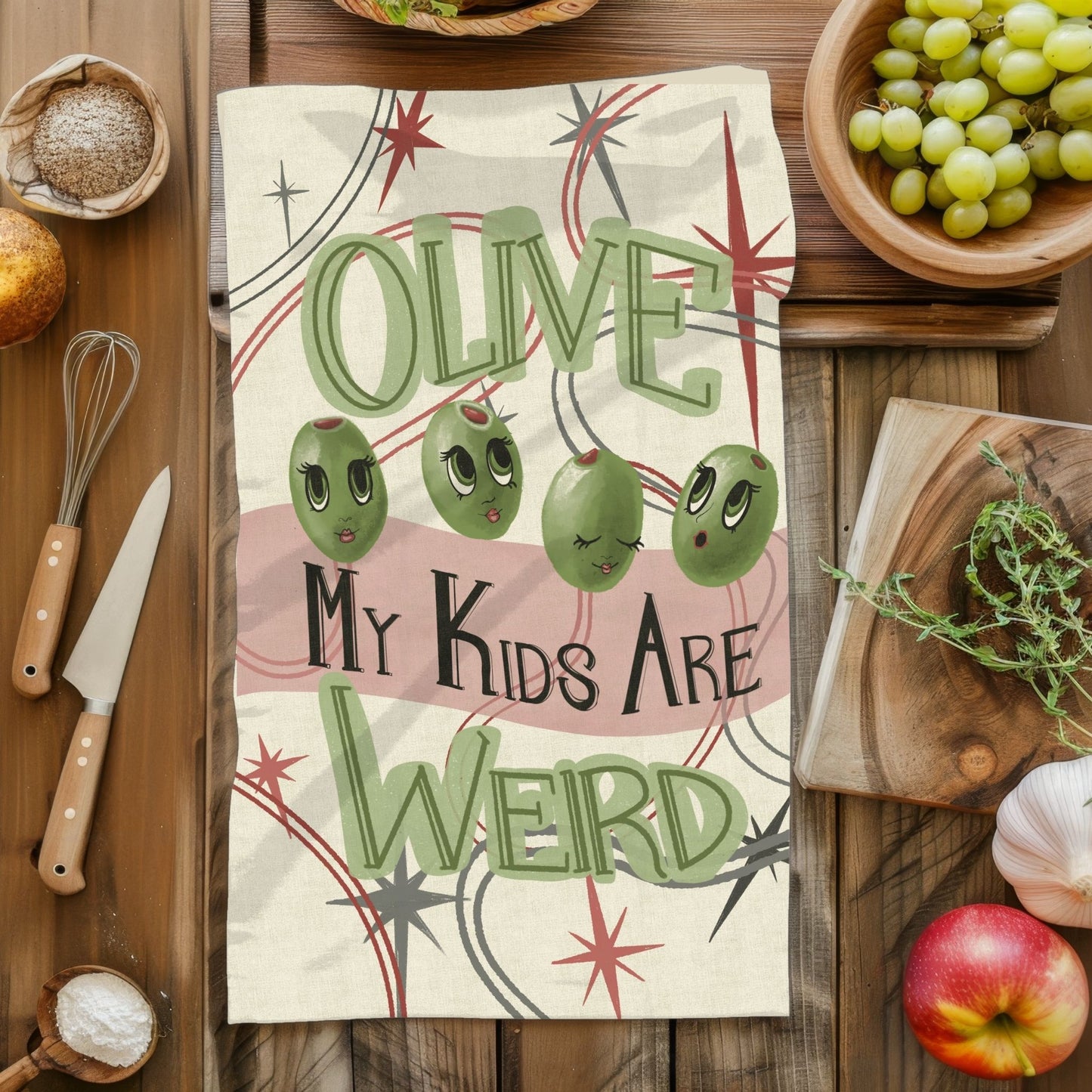 Retro mid-century kitchen tea towel with olive pun illustration laid flat on wooden surface surrounded by cooking props, original artwork on cotton twill