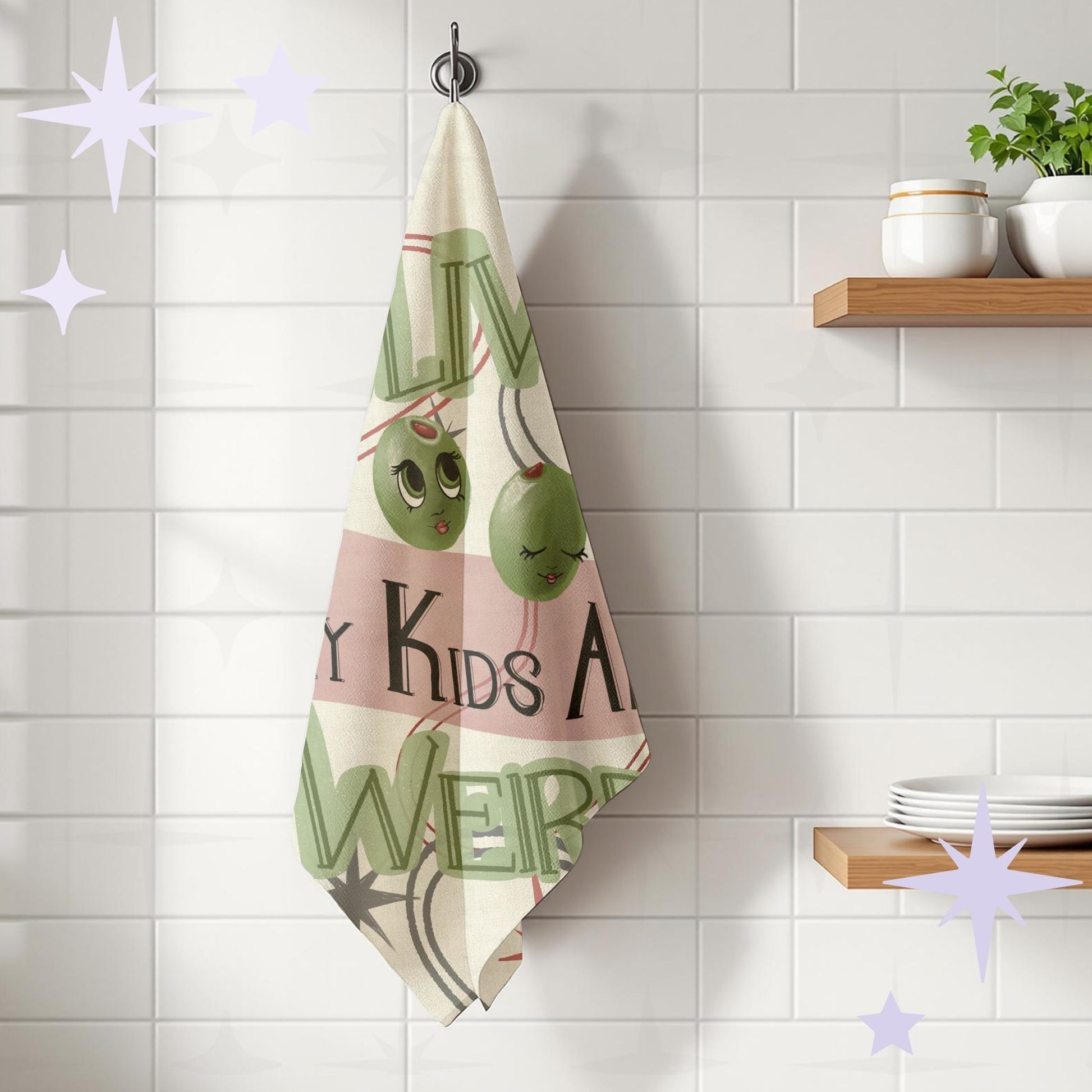 Olive pun tea towel hanging on hook in white kitchen with subway tile, showing cartoon olive faces and retro hand-lettered typography, cotton twill