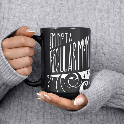 Person in grey cable-knit sweater holding black goth mom mug showing I'm Not a Regular Mom hand lettering in gothic style, 15oz ceramic