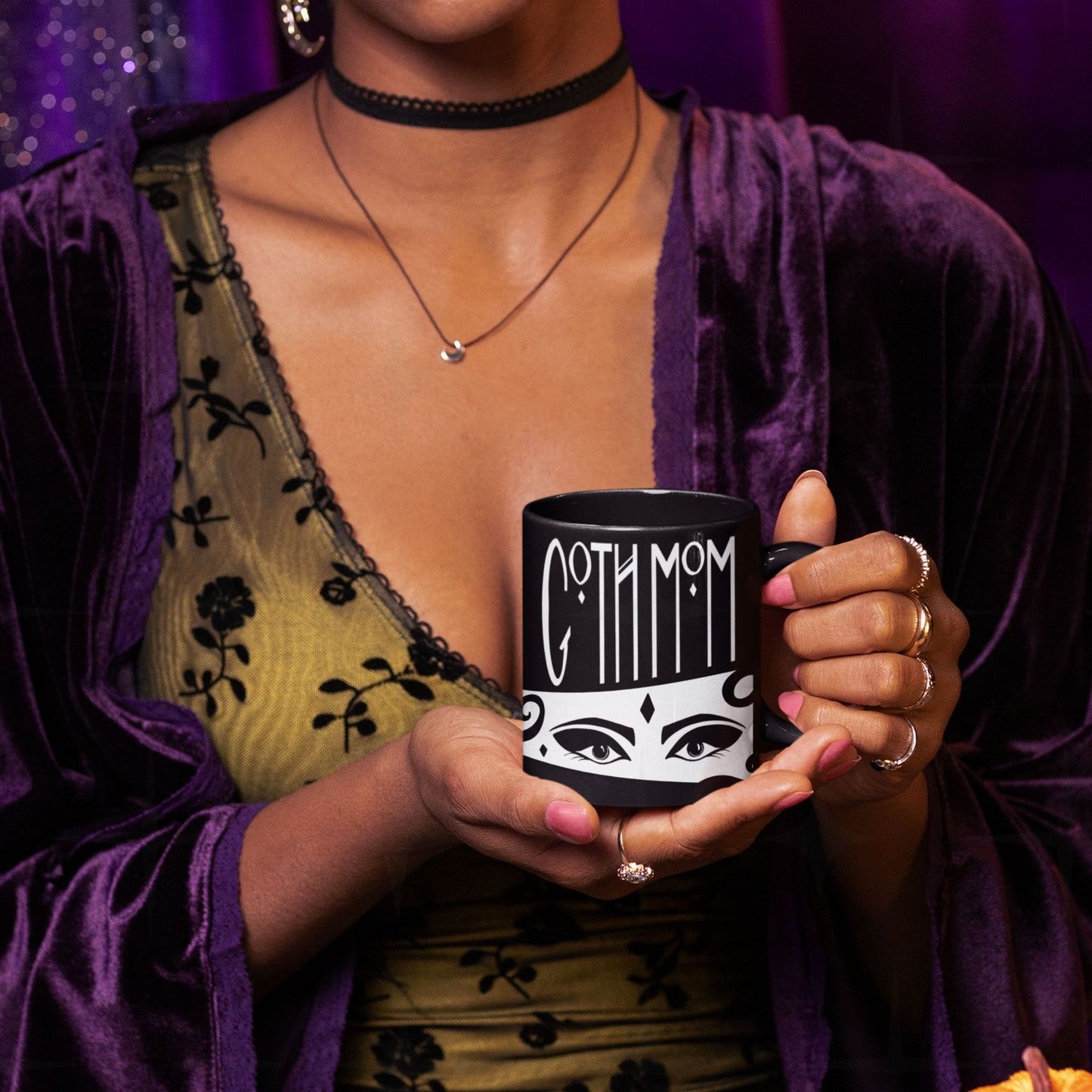 Woman in purple velvet robe and gold lace top holding black goth mom mug with Goth Mom lettering and illustrated eyes, styled gothic aesthetic shoot