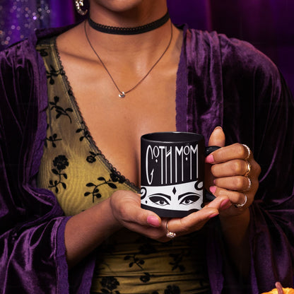 Woman in purple velvet robe and gold lace top holding black goth mom mug with Goth Mom lettering and illustrated eyes, styled gothic aesthetic shoot