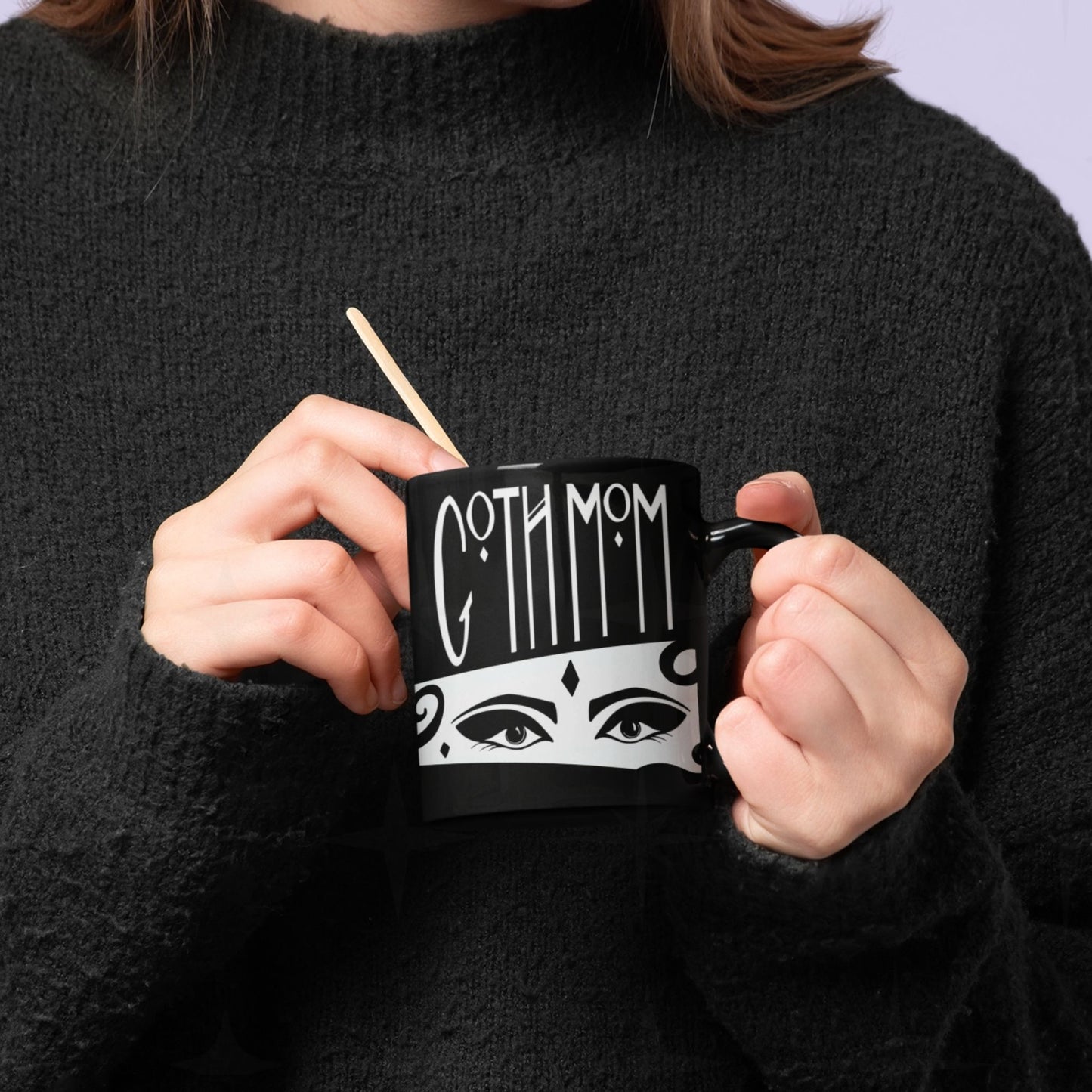 Hands holding black ceramic goth mom mug with hand-lettered gothic typography and illustrated masquerade eyes, wooden stirrer resting in cup