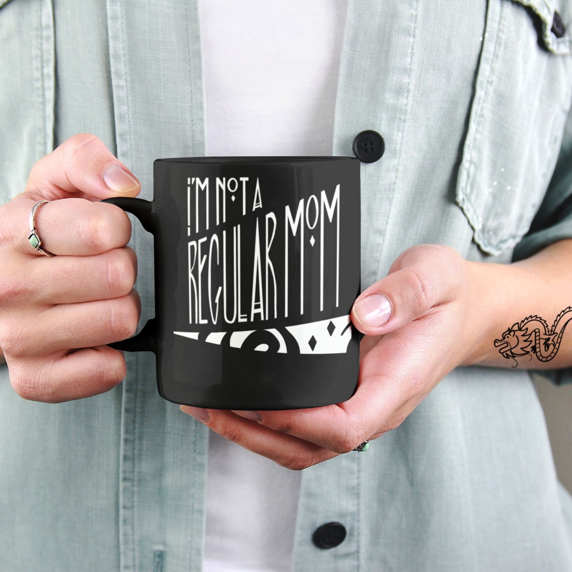 Tattooed hands holding black ceramic goth mom coffee mug with gothic hand lettering reading I'm Not a Regular Mom, 11oz size