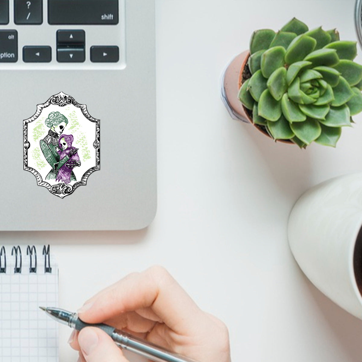 Gothic skeleton sticker applied to a silver laptop lid beside a succulent and a notepad