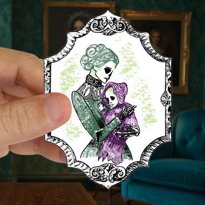 Hand holding a die cut skeleton sticker of a Victorian mother and baby against a green wall of gilt framed portraits
