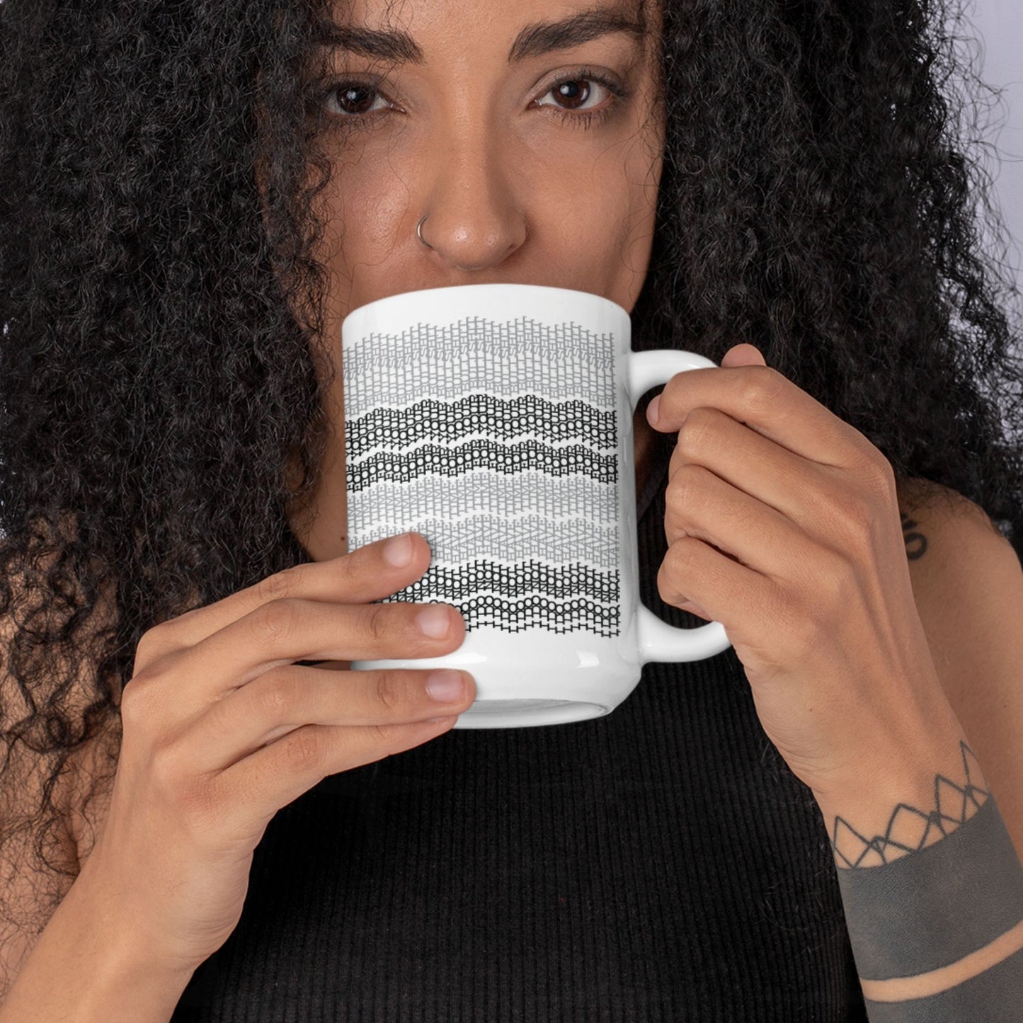 Woman with curly hair and nose ring drinking from white ceramic hidden message mug with gray and black embroidery stripe pattern, arm tattoo visible