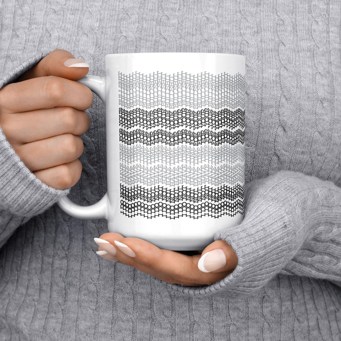 White ceramic hidden message mug with dense embroidery-style gray stripe pattern held in two hands against a gray cable-knit sweater