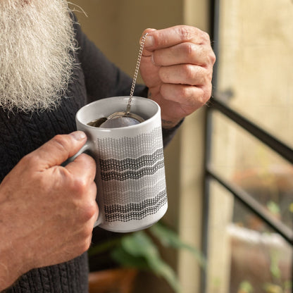 Person with white beard and tattoos brewing loose leaf tea in gray stripe embroidery pattern ceramic mug using a metal tea ball strainer