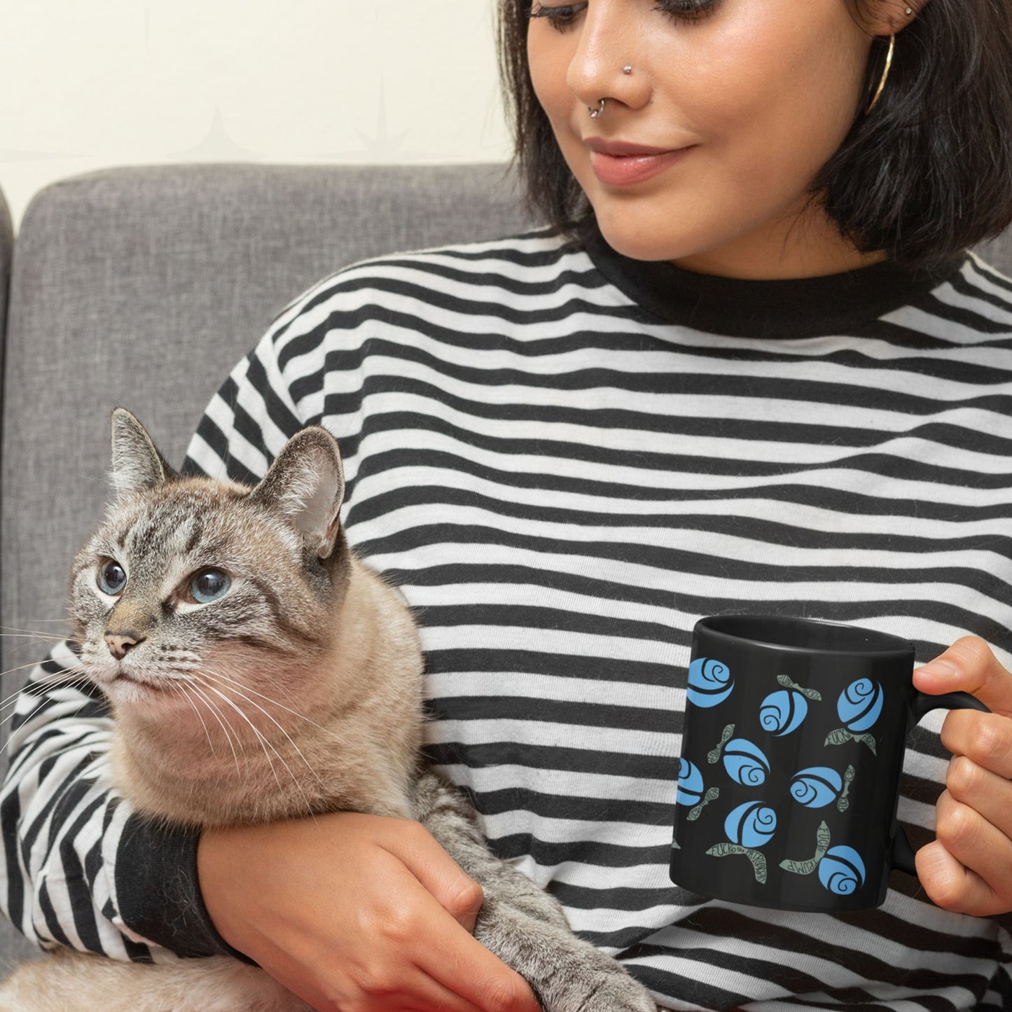 Woman holding blue hidden message protest mug on black ceramic while seated with a grey cat, floral rose pattern with subversive hand-lettered leaves visible