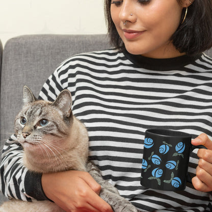 Woman holding blue hidden message protest mug on black ceramic while seated with a grey cat, floral rose pattern with subversive hand-lettered leaves visible