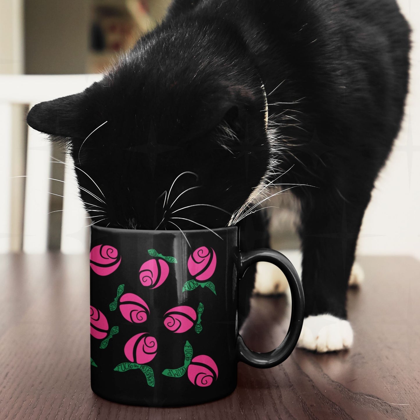 Black and white cat nosing into hot pink hidden message protest mug on wooden surface, graphic rose pattern with green hand-lettered leaves on black ceramic