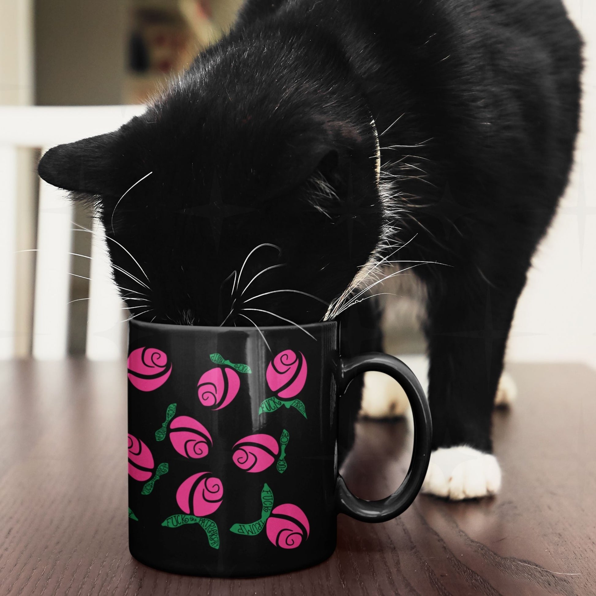 Black and white cat nosing into hot pink hidden message protest mug on wooden surface, graphic rose pattern with green hand-lettered leaves on black ceramic