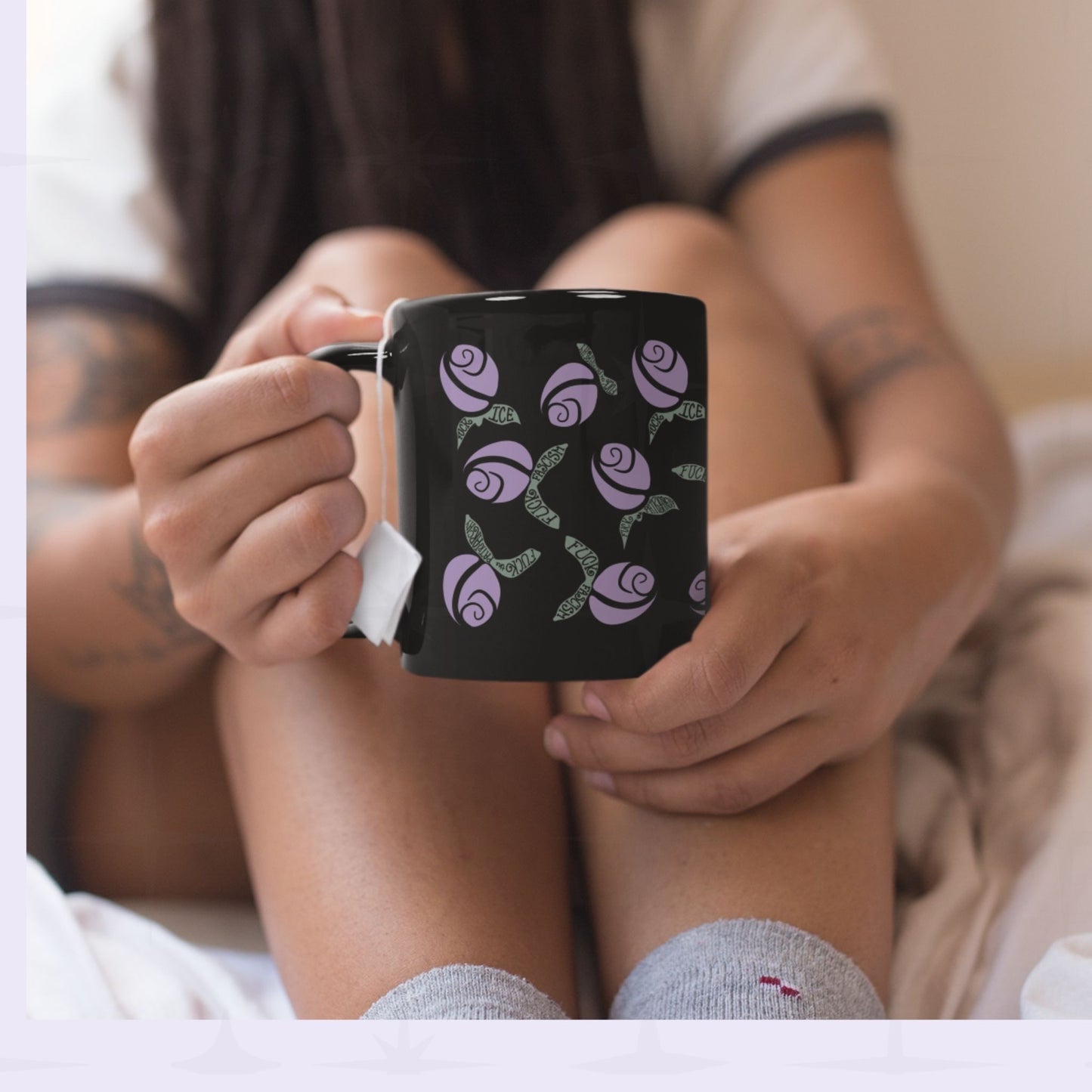 Person in pajamas holding lilac purple hidden message protest mug with a tea bag, graphic rosebuds and subversive hand-lettered sage green leaves clearly visible on black ceramic
