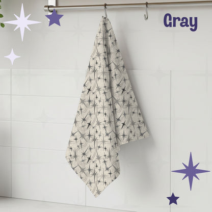 Warm gray atomic starburst mid century modern tea towel hanging from a chrome hook against white subway tile, showing full pattern of charcoal and gray starburst motifs on a natural oatmeal cotton twill background