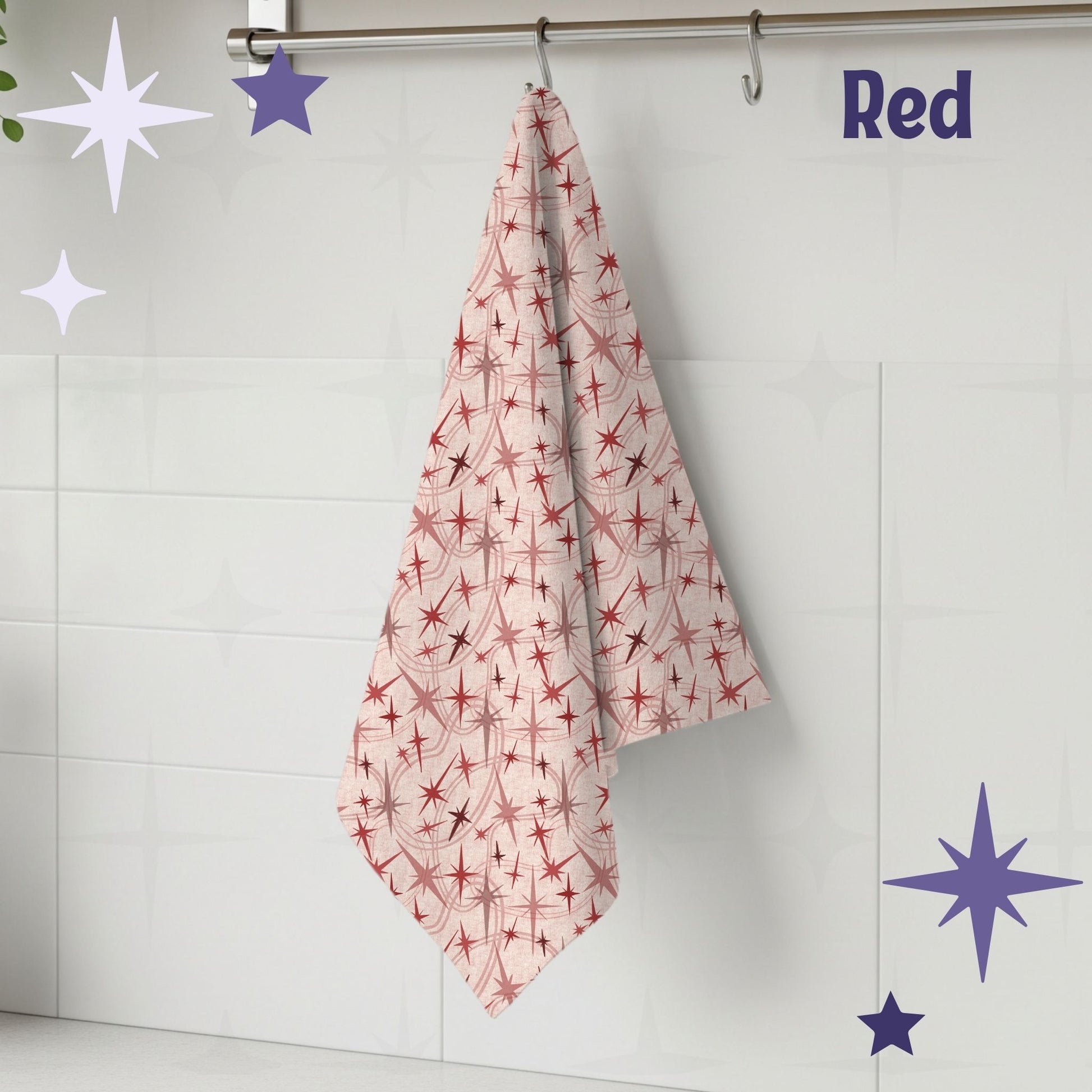 Red and dusty rose atomic starburst mid century modern tea towel hanging from a chrome hook against white subway tile, showing full pattern of crimson and blush starburst motifs on a nude cream cotton twill background