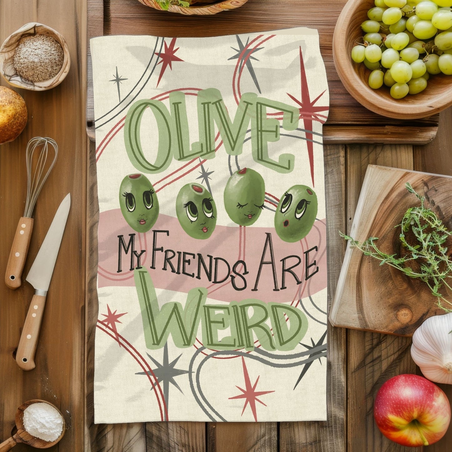 Retro mid-century olive pun tea towel laid flat on a rustic wooden kitchen surface surrounded by produce and baking tools