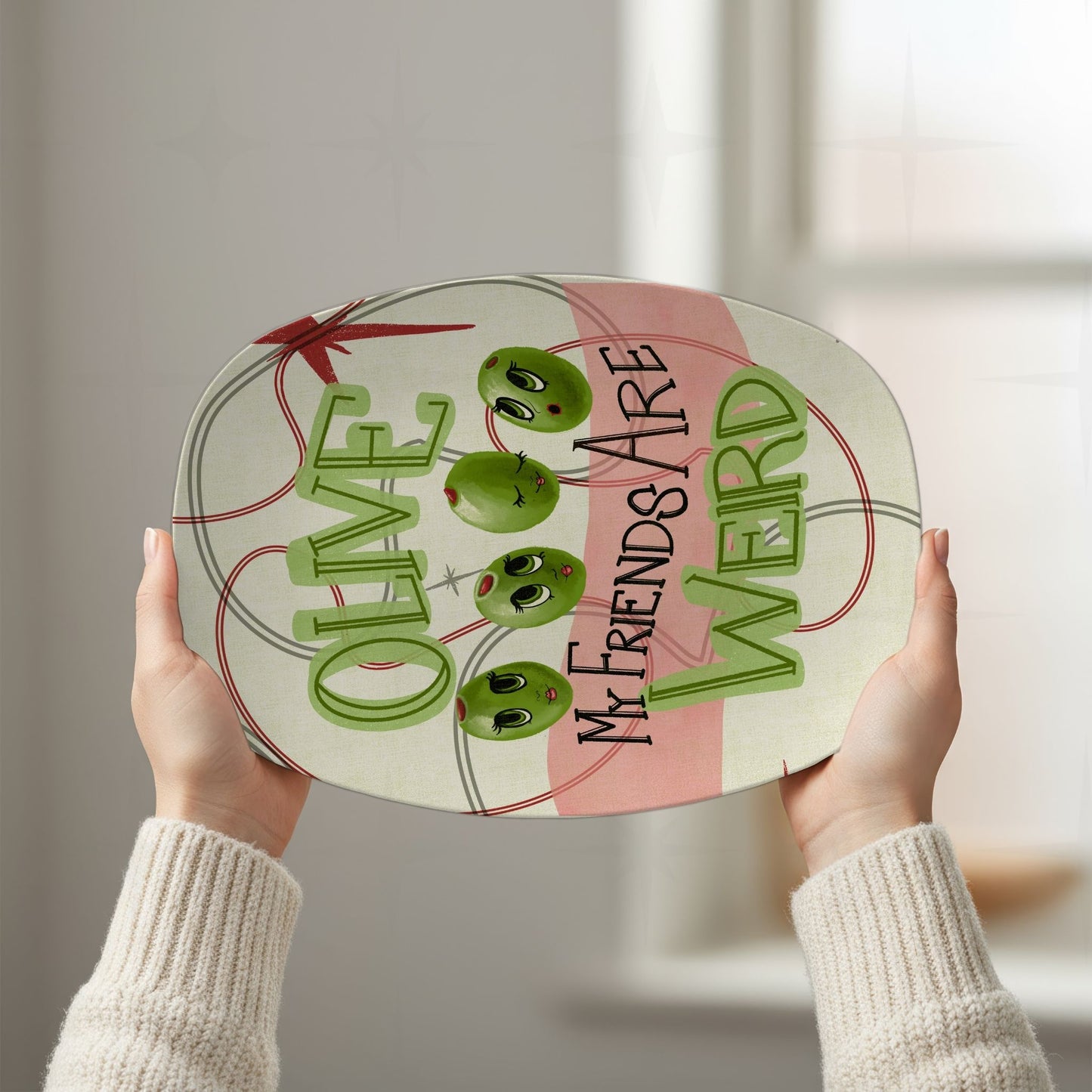 Two hands holding retro olive pun platter overhead against bright white background, showing full "Olive my friends are weird" mid-century illustrated design with atomic starburst motif and oval line framing