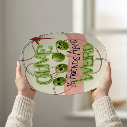 Two hands holding retro olive pun platter overhead against bright white background, showing full "Olive my friends are weird" mid-century illustrated design with atomic starburst motif and oval line framing