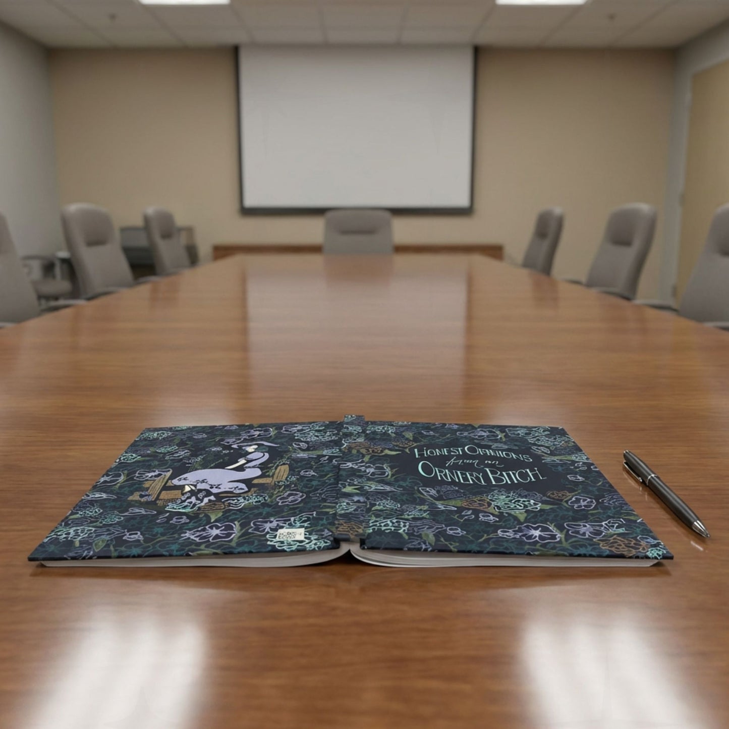 Ornery Bitch sweary journal open flat on conference room table beside a pen, showing full wrap dark floral and cowgirl pinup artwork on both covers