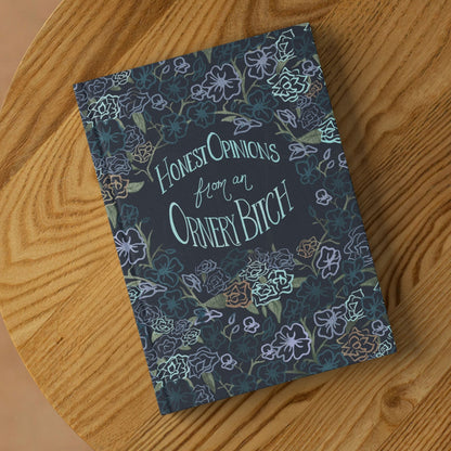 Sweary dark floral hardcover journal lying on round wooden surface showing front cover with hand-lettered Honest Opinions from an Ornery Bitch text and gothic botanical pattern