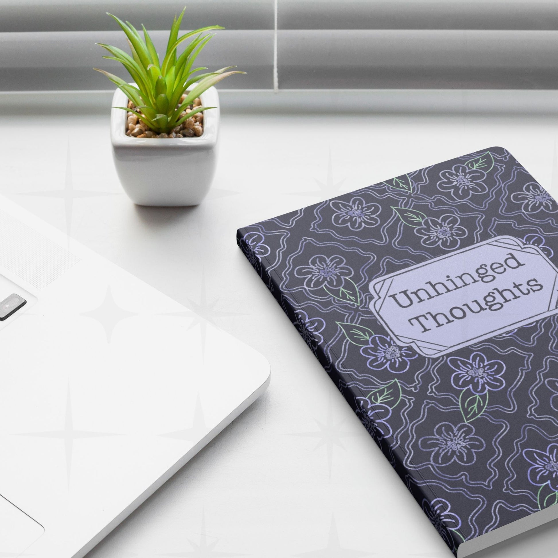 Unhinged Thoughts dark floral softcover journal placed on a desk next to a laptop and a plant
