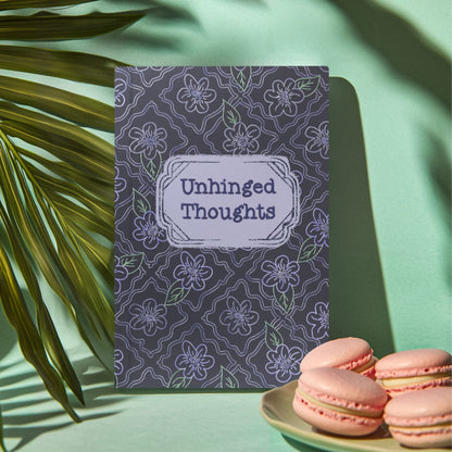 Unhinged Thoughts dark floral softcover journal standing upright beside pink macarons and palm frond on mint green background