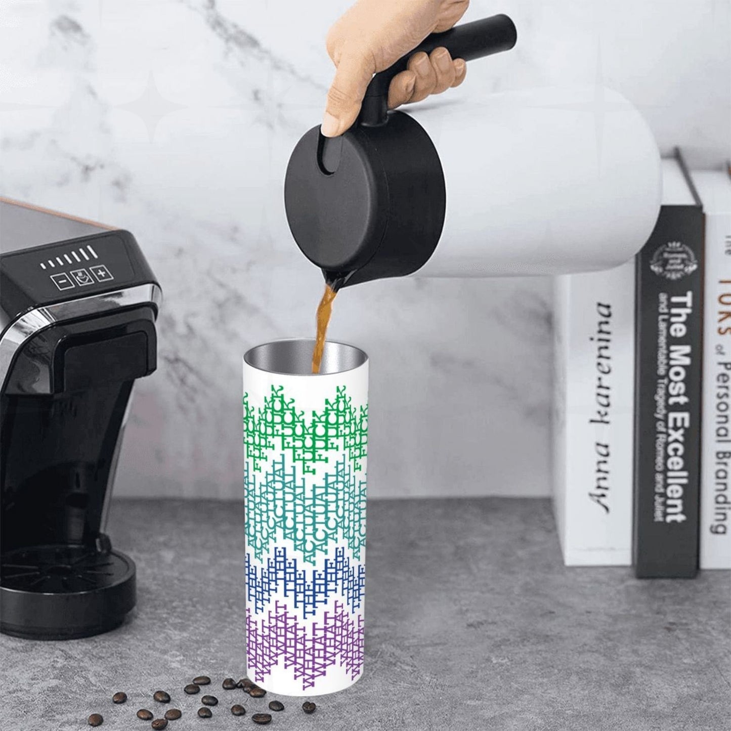Coffee being poured into the bright stripes What The Actual Fuck hidden message tumbler on a kitchen counter beside an espresso machine and stacked books
